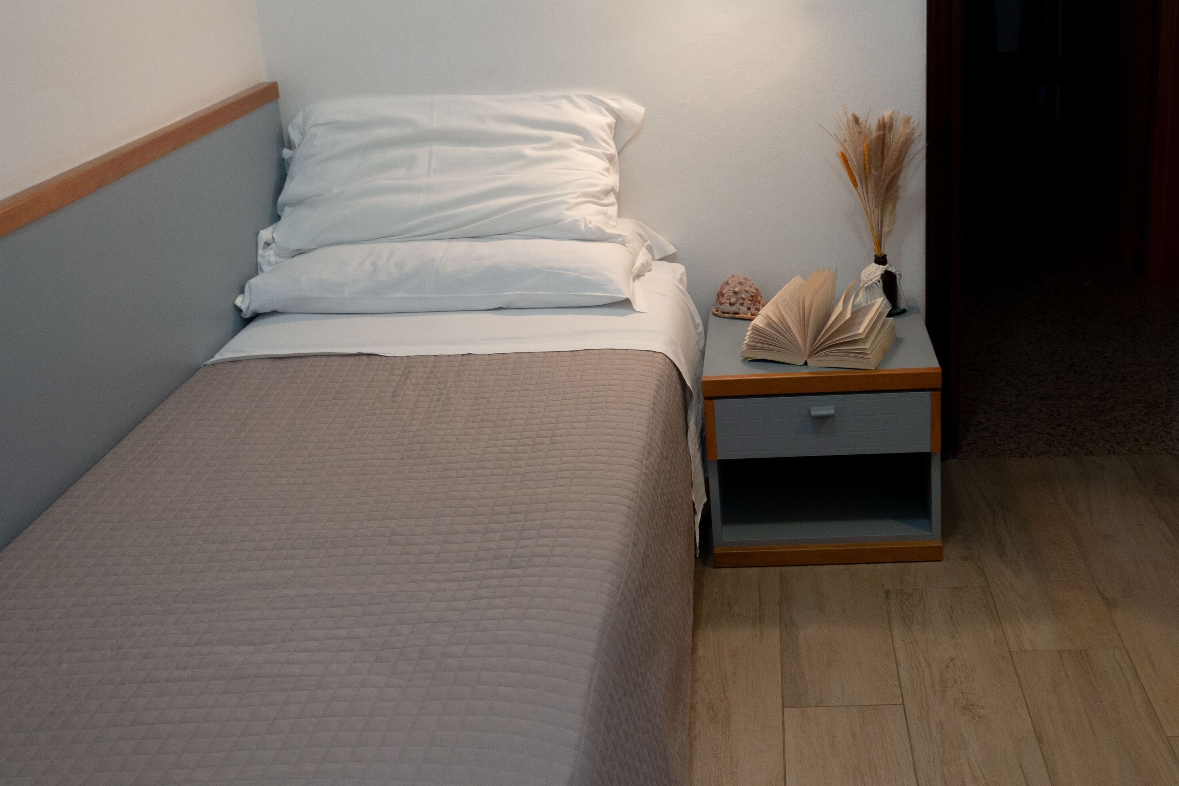 A neatly made bed with a quilted cover, a bedside table with a vase of dried flowers, and a small drawer.