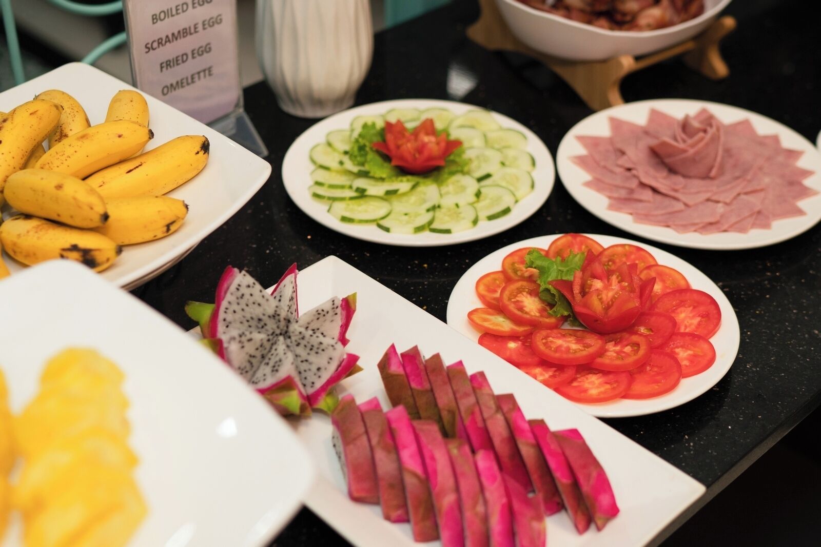 A variety of food items on a table, including bananas, cucumber salad, tomato salad, and sliced meat.