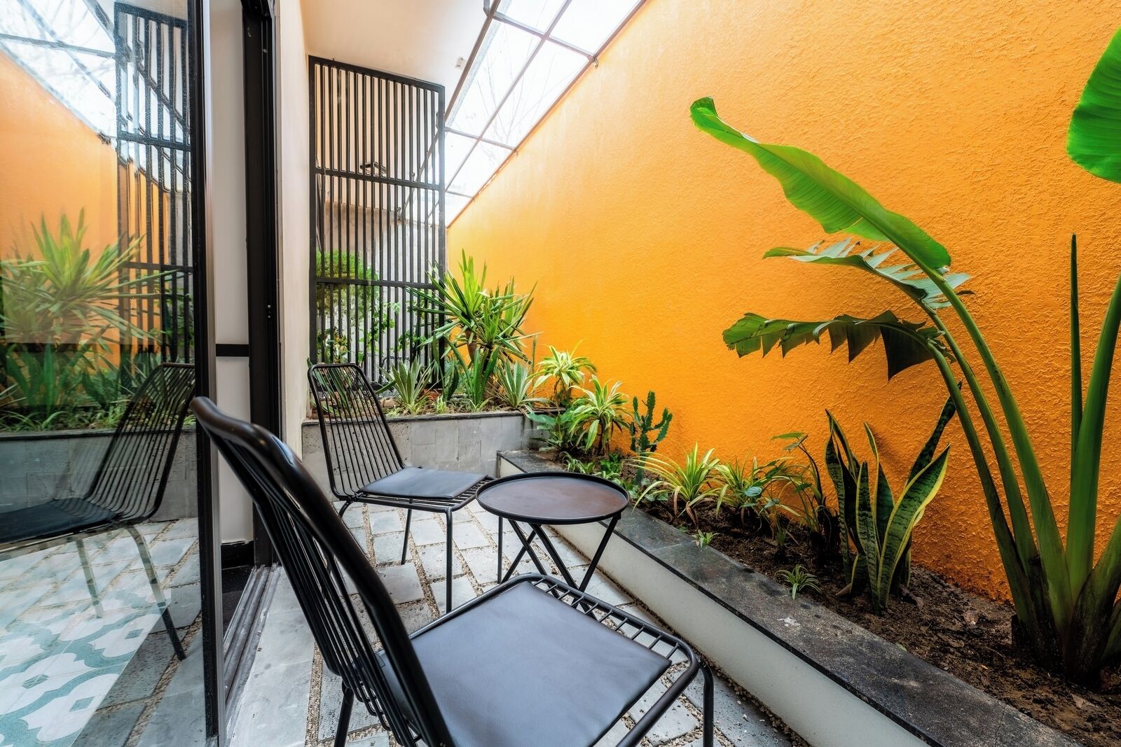 A courtyard with a tiled floor, a black metal chair, a small round table, and various plants.