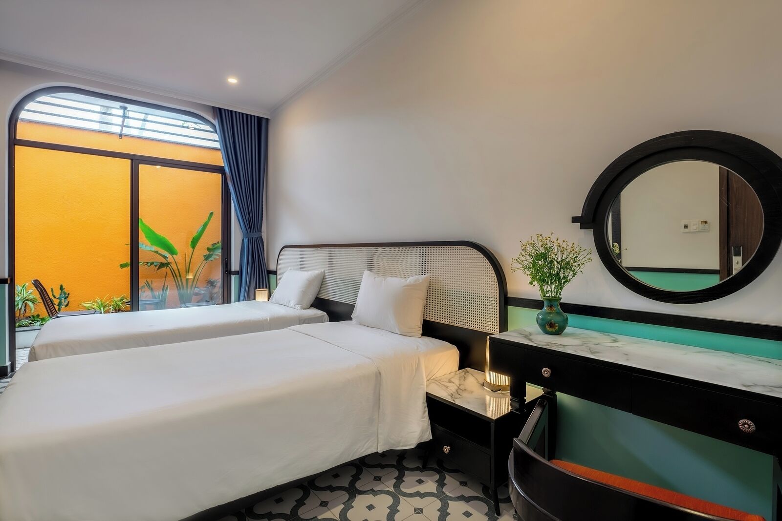 A hotel room with two beds, a large window with curtains, a bedside table with a vase, and a round mirror.