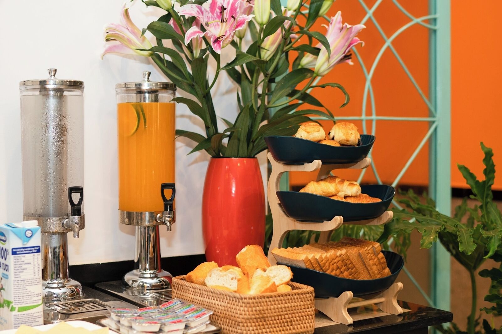 A breakfast setup with a juice dispenser, a vase of flowers, and a tiered serving tray with pastries and bread.