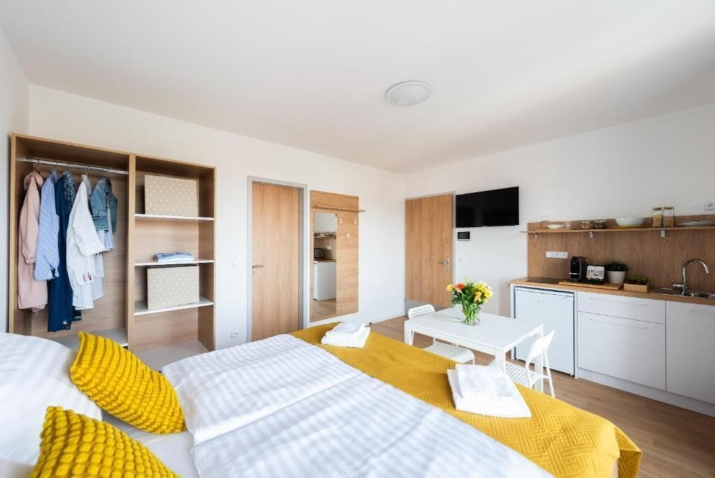 A modern hotel room with a bed, a dining table with chairs, a small kitchen area, and a walk-in closet.