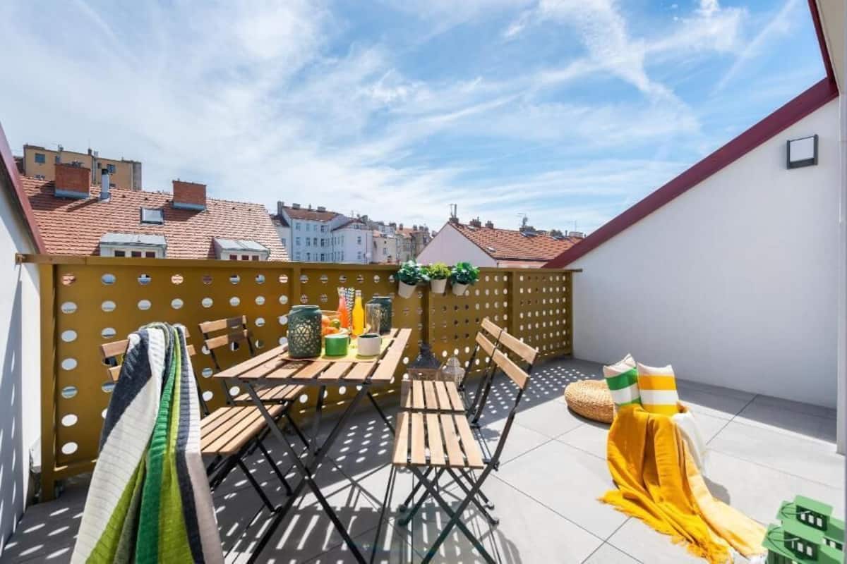 Une terrasse sur le toit, avec des meubles en bois, une nappe jaune et une vue sur les bâtiments en arrière-plan.