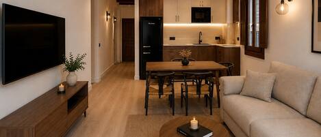 A modern living room with a flat-screen TV, a wooden coffee table with a book and candle, a beige sofa, and a kitchen in the background.