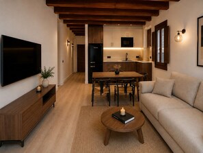 Una sala moderna con televisor de pantalla plana, una mesa de centro de madera con un libro y una vela, un sofá color beige y una cocina al fondo.