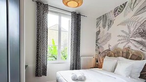Et soveværelse med en seng, et vindue med gardiner, en væg med bladmotiver og en hængende lampe.