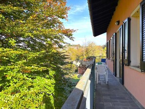 A balcony with a view of a garden, a chair, and a building with shutters. - Lakeview Apartment in Luino for 4 (Luino)