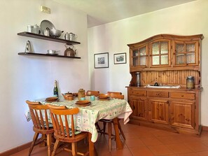 A dining area with a table set for four, surrounded by wooden chairs. A wooden cabinet with glass doors is to the right, and a shelf above the table holds kitchen utensils. - Lakeview Apartment in Luino for 4 (Luino)