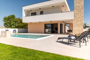 Una casa moderna de dos pisos con piscina, mobiliario exterior y un pilar de piedra.