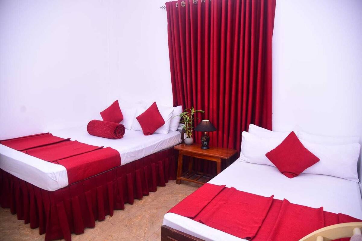 A room with two beds, each with red bedding and pillows, a small table with a lamp, and a curtain.