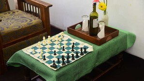 A chessboard on a table with a green tablecloth, a wooden chair with a patterned cushion, and a tray with a bottle, glasses, and flowers. - Travel Rest Inn (Ella)