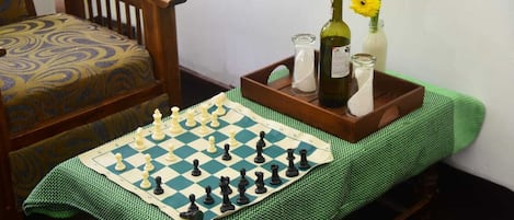 A chessboard on a table with a green tablecloth, a wooden chair with a patterned cushion, and a tray with a bottle, glasses, and flowers.