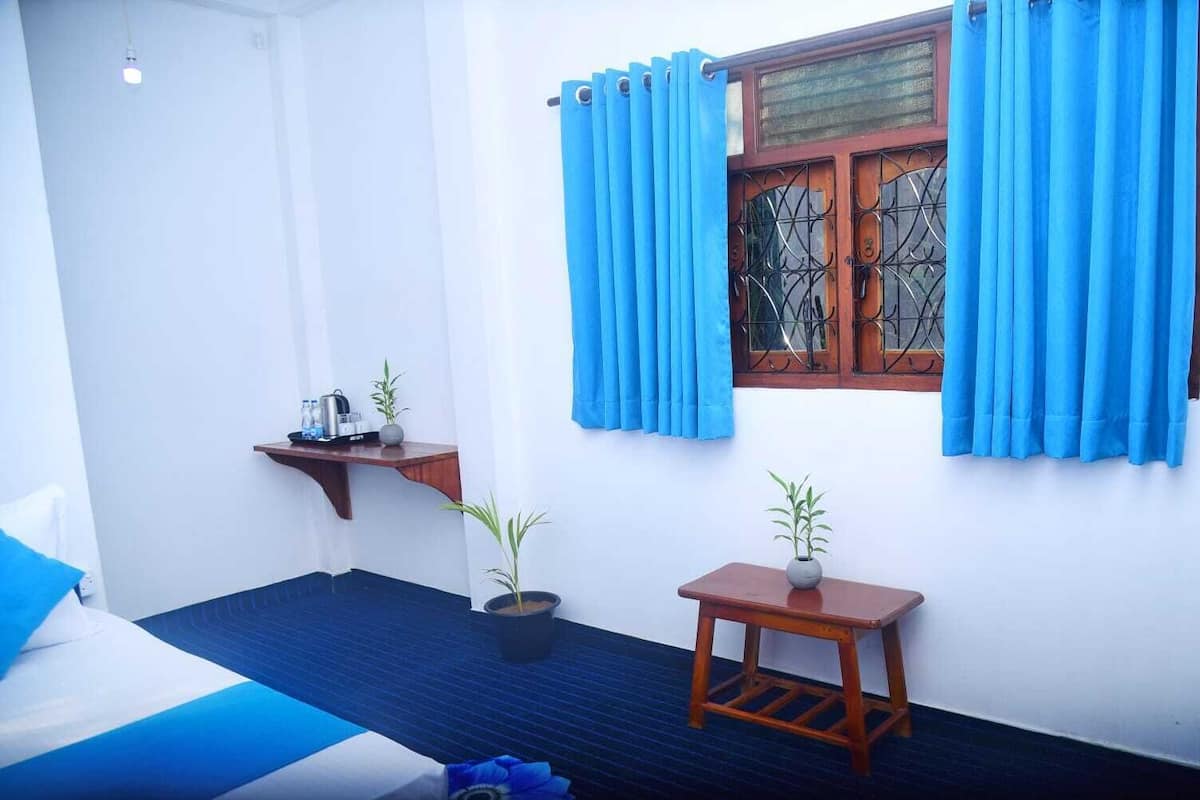 A room with a bed, a small table with a plant, a shelf with items, and a window with curtains.