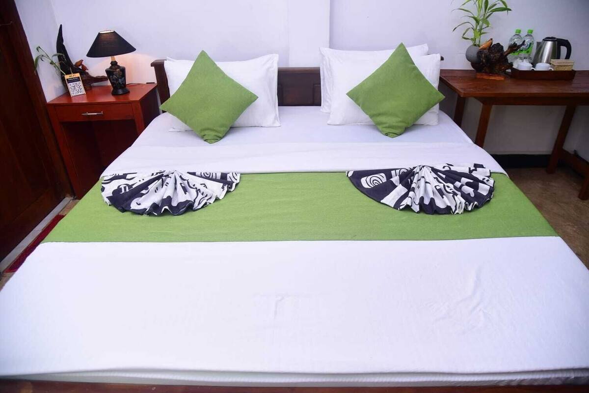 A neatly made bed with a green throw blanket and two pillows, a bedside table with a lamp and a small plant, and a nightstand with a decorative item.