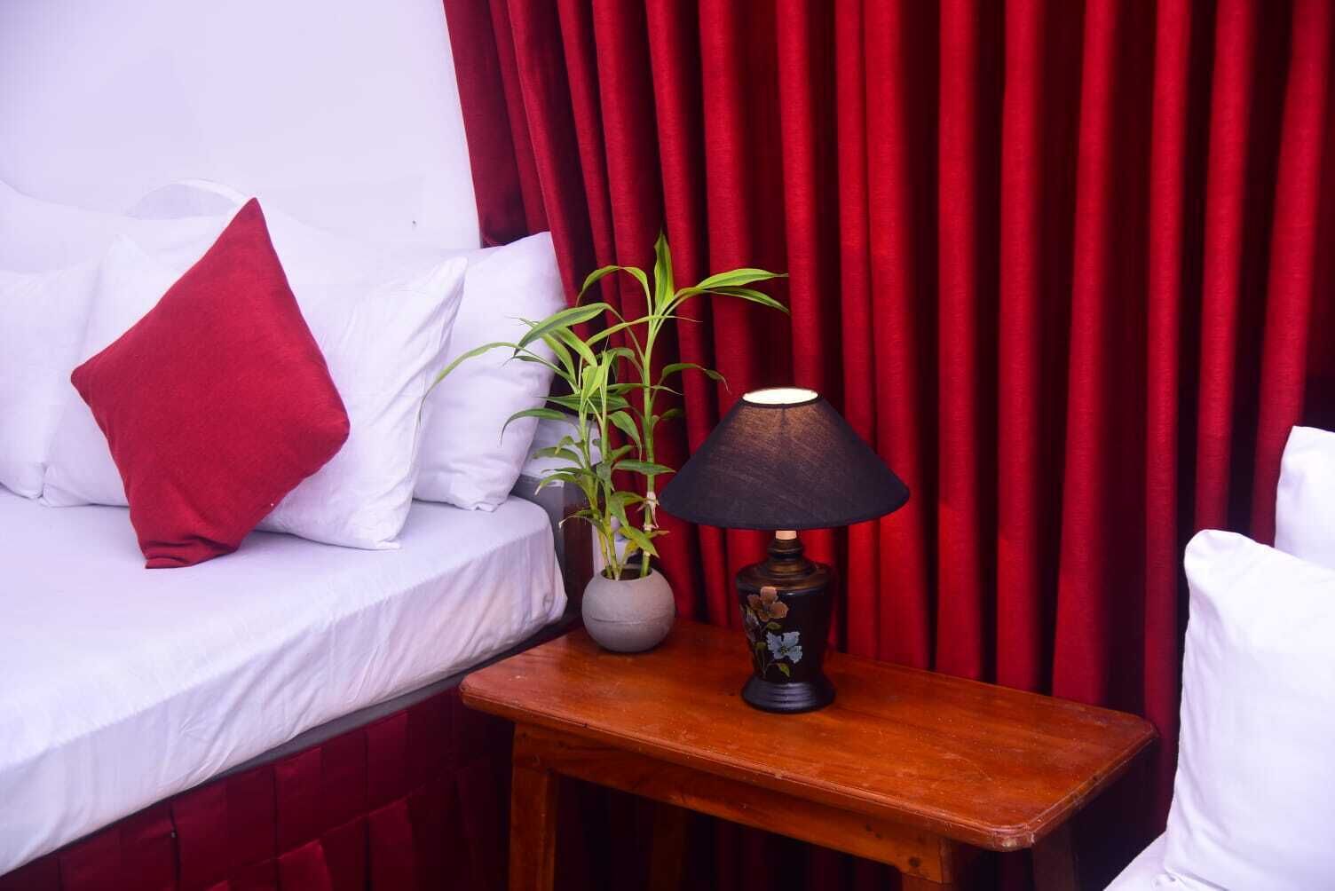 Un lit avec une literie blanche et des oreillers rouges, une petite table avec une lampe et une plante en pot, et des rideaux rouges en arrière-plan.