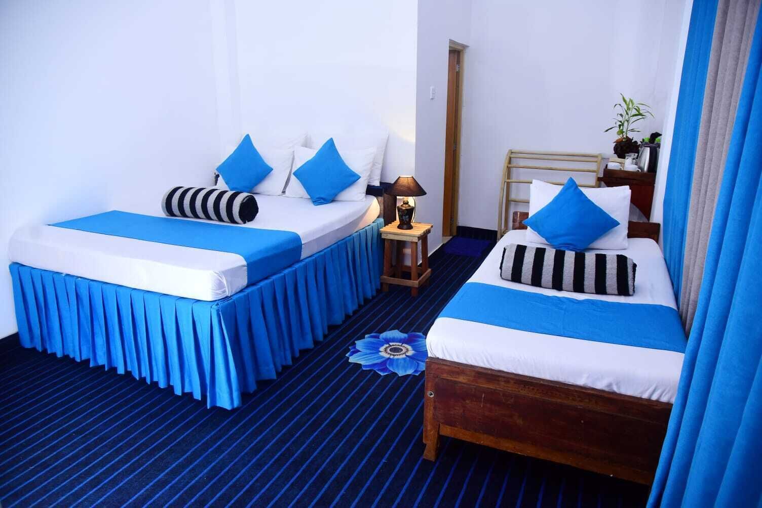 Habitación de hotel con dos camas, cada una con ropa de cama y almohadas azules, una mesita con lámpara y un banco de madera.