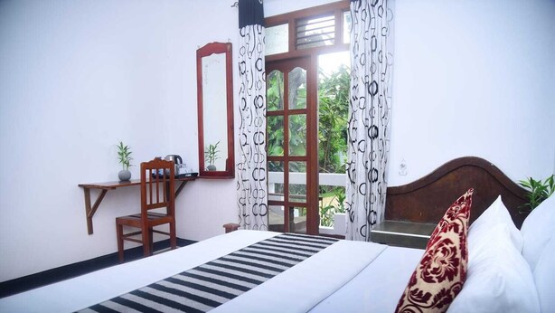 A bedroom with a bed, a desk, a chair, a window with curtains, and a view of greenery outside. - Travel Rest Inn (Ella)