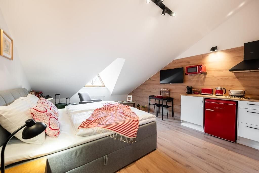 Habitación compacta y moderna con cama doble alta, kitchenette y televisor montado en la pared.