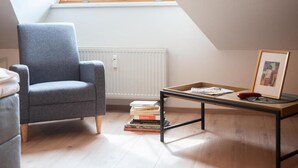 Una habitación con techo inclinado, un sillón gris, una mesa de centro de madera con libros y un cuadro enmarcado, y un radiador.