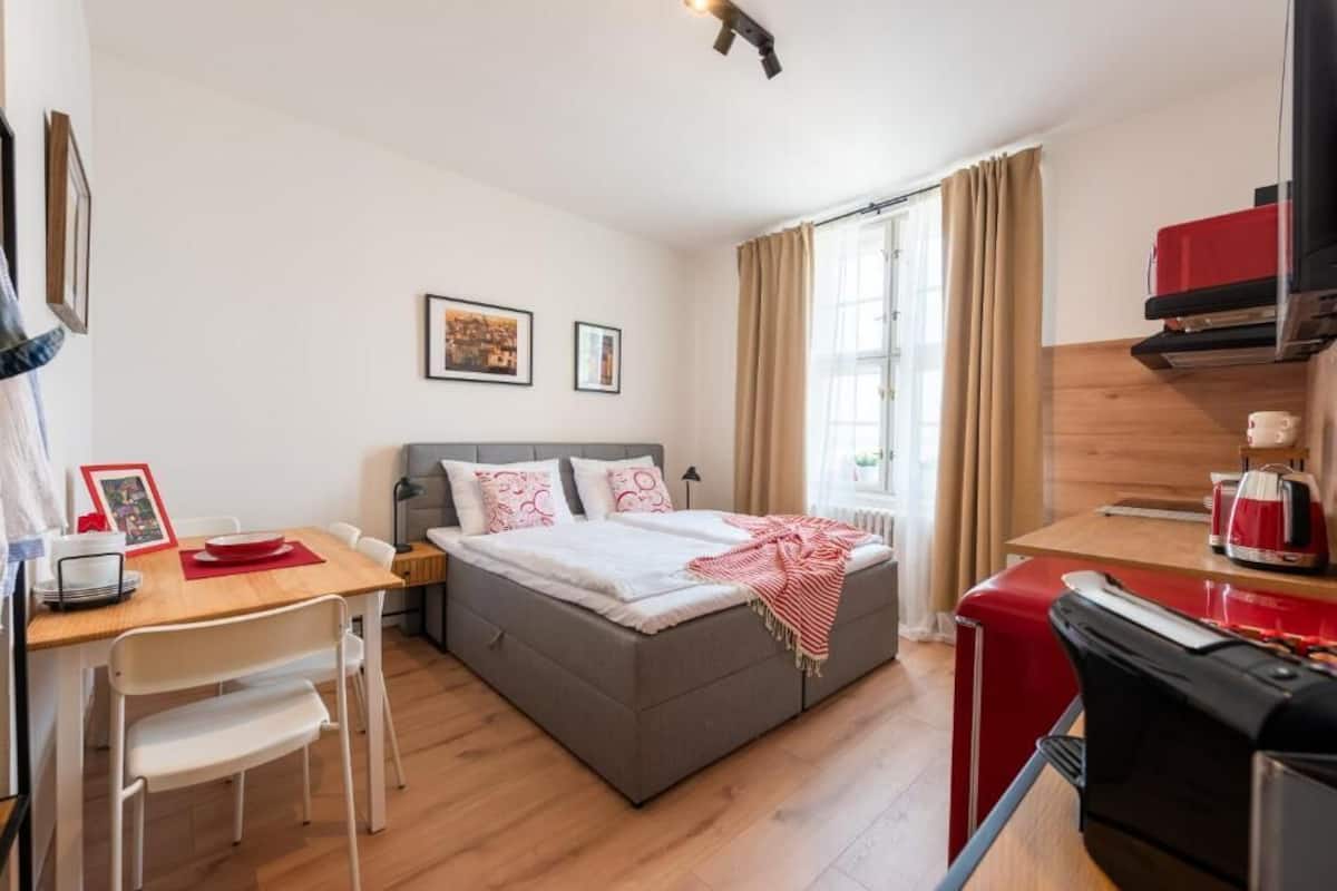 Ein kleines Zimmer mit Bett, Schreibtisch, Stuhl, einer Küchenzeile und einem Fenster mit Vorhängen.