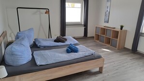 A modern bedroom with a wooden bed, patterned pillows, a bedside table, a floor lamp, a window with curtains, and a small plant. - Apartment 'Für 4 Personen 65 M²' with Private Terrace, Private Garden and Wi-Fi (Dornum)