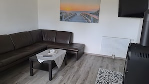 A living room with a brown leather sofa, a small table with a remote control, a radiator, and a TV mounted on the wall. - Apartment 'Für 4 Personen 65 M²' with Private Terrace, Private Garden and Wi-Fi (Dornum)