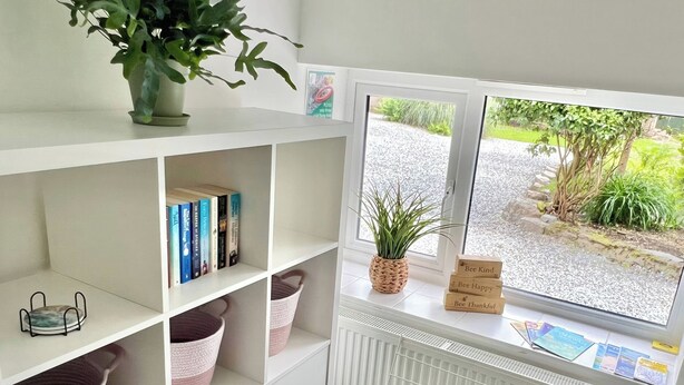 A room with a white radiator, a window with a view of greenery, a bookshelf with books and baskets, and a plant on the windowsill. - Hydrangea Cottage (St Austell)