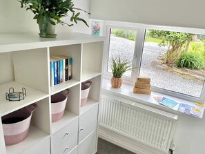 Una habitación con un radiador blanco, una ventana con vistas a zonas verdes, una estantería con libros y cestas, y una planta en el alféizar.