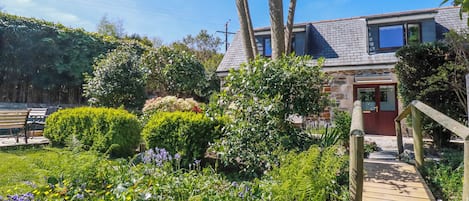 Un jardin avec une terrasse en bois, un bâtiment en pierre doté d’une porte rouge et un banc.
