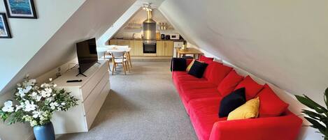 An attic-style room with a red sofa, a TV, a dining table, and a kitchen area.