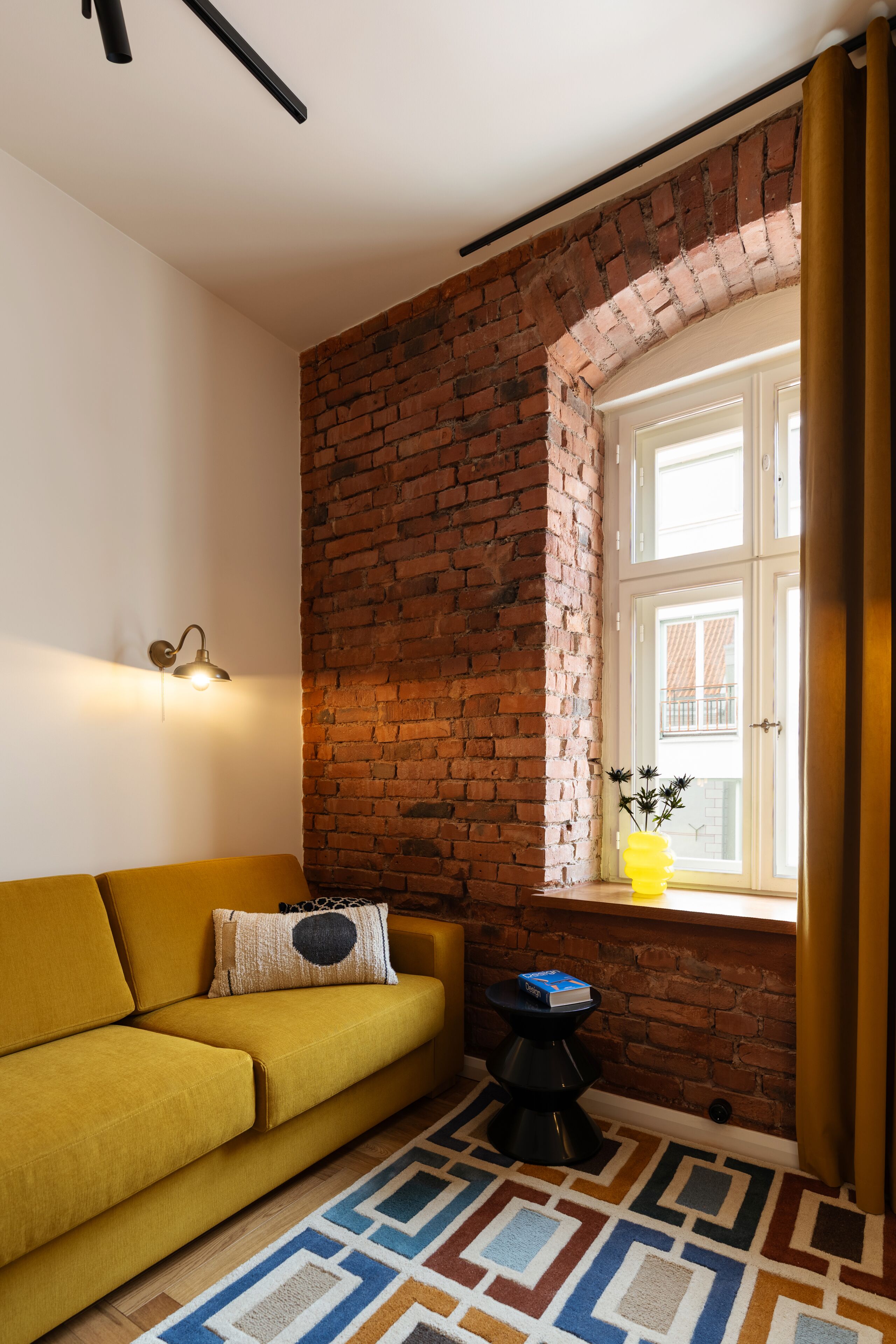 A room with a yellow sofa, a brick wall, a window with curtains, a small table with a book, and a patterned rug.