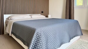 Un lit recouvert d’une courtepointe grise matelassée, une tête de lit en lattes de bois, une table de chevet blanche avec une lampe, et une vue sur la verdure par la fenêtre.