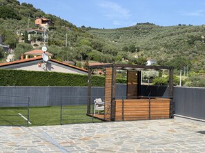 Un gazebo con pavimento in pietra, circondato da una recinzione, con case e colline sullo sfondo.