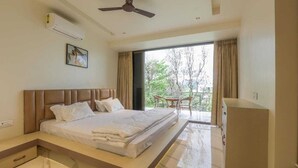 A hotel room with a bed, a ceiling fan, an air conditioning unit, a window with curtains, and a view of trees outside.