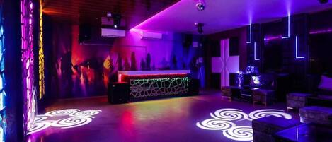 A modern bar area with a neon-lit reception desk, ambient lighting, and a patterned floor design.