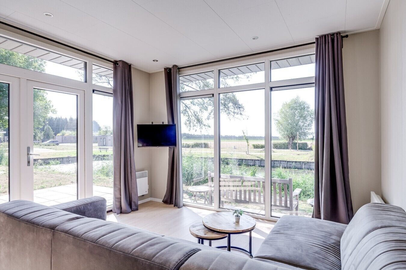 A modern living room with a large TV, grey sofas, and a view of a garden and outdoor seating area.