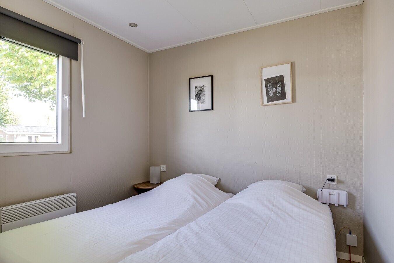 A bedroom with two beds, a window with a blind, a radiator, and two framed pictures on the wall.
