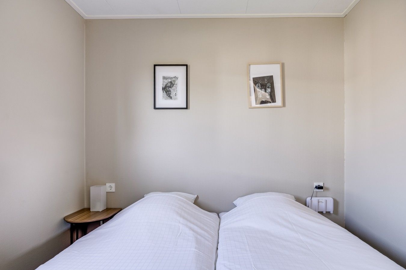 Et hotelværelse med to senge, et lille trænatbord og to billeder i rammer på væggen.