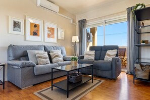 A living room with a grey sofa, a matching armchair, a glass coffee table, and a floor lamp. There are framed artworks on the wall, a window with curtains, and a balcony with a railing. - Charming Apartment for 4 Near Golf & Sea (Rincón de la Victoria)