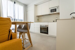 A modern kitchen with white cabinets, a microwave, and an oven. There is a wooden chair and a small table. - Brand New · Modern Comfort in Málaga by VGC (Málaga)