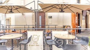 Un café al aire libre con mesas, sillas, sombrillas y un edificio con una puerta y ventanas al fondo.