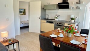 Eine kompakte Küche mit modernen Geräten, ein Essbereich mit Tisch für zwei Personen und ein kleines Schlafzimmer, das durch eine offene Tür sichtbar ist.