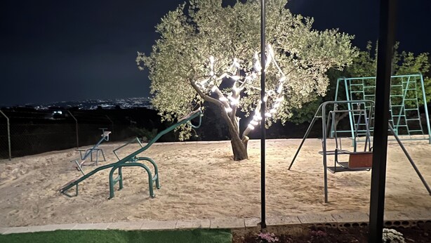 Une aire de jeux avec des balançoires et une structure d’escalade, entourée de sable et d’un arbre.