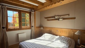A bedroom with a wooden ceiling, a bed with checkered bedding, a window with curtains, and a mounted ski rack.