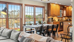 A spacious kitchen with wooden cabinets, a dining area with a table and chairs, and large windows overlooking a natural landscape. - Sleeps 20! 2 Lakefront Homes w/ Hot Tub, Firepit, Game Room, Community Amenities (Long Pond)