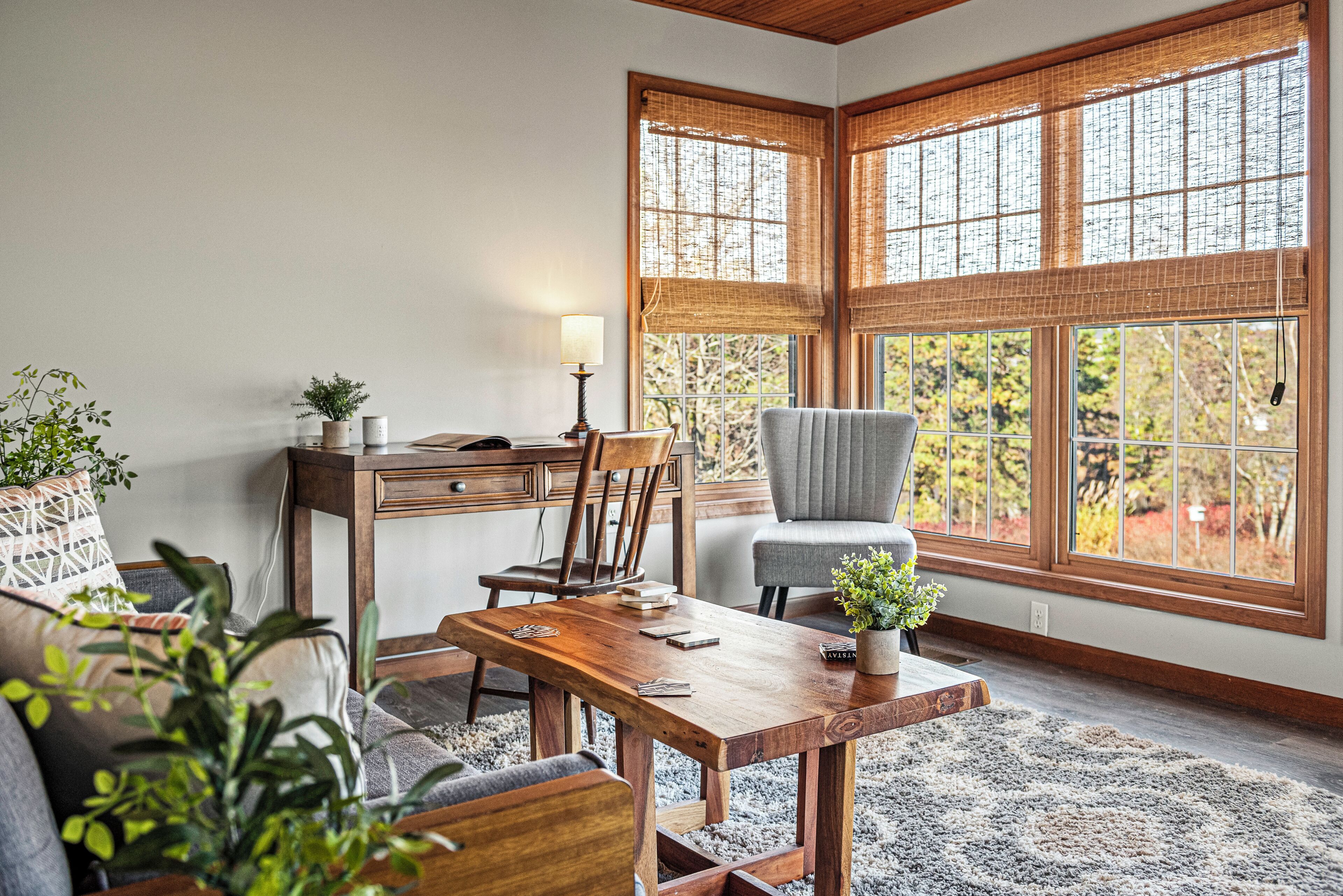 Una habitación acogedora con una mesa de comedor de madera, una silla cómoda, un sofá y un escritorio con una lámpara. Amplias ventanas con persianas de bambú dejan entrar la luz natural.
