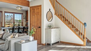 A spacious living room with a wooden staircase, a dining area with a large window, and a cozy seating area with a sofa and a coffee table. - Sleeps 20! 2 Lakefront Homes w/ Hot Tub, Firepit, Game Room, Community Amenities (Long Pond)