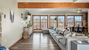 A spacious living room with a large sectional sofa, wooden beams on the ceiling, and large windows offering a view of the outdoors. - Sleeps 20! 2 Lakefront Homes w/ Hot Tub, Firepit, Game Room, Community Amenities (Long Pond)
