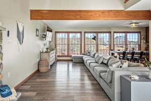 Ein großzügiges Wohnzimmer mit einer großen Eckcouch, Holzbalken an der Decke und großen Fenstern mit Blick ins Freie.