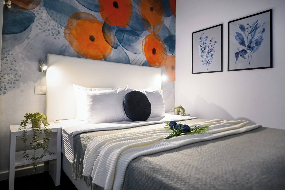 Un dormitorio con cama, mesitas de noche, una mesita con una planta y un cabecero con dos cuadros.